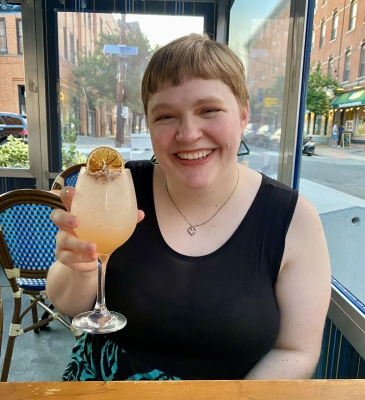 Melody is sitting a table, wearing black dress and green triangle earrings, holding a cocktail and smiling.